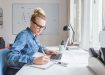 Blonde woman wearing jeans shirt and nerd glasses sitting at the desk in an office and working on laptop.