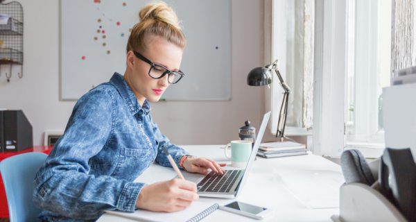 Blonde woman wearing jeans shirt and nerd glasses sitting at the desk in an office and working on laptop.